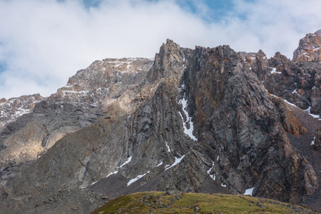 Atmospheric landscape with sharp pinnacle on sunlit high mountain range under clouds in blue sky. Beautiful alpine scenery with peaked mountain top in sunlight under cloudy sky at changeable weather.の写真素材