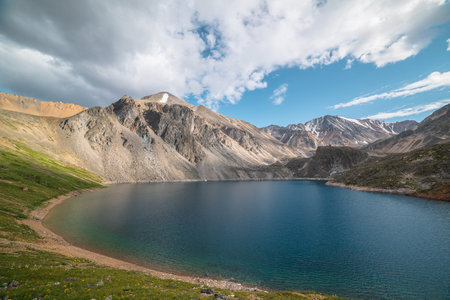 Deep mountain lake of phantom blue color among high mountains with pointed peak in changeable weather. Wonderful dramatic view to deep blue mountain lake among sunlit sharp rocks under cloudy sky.の写真素材