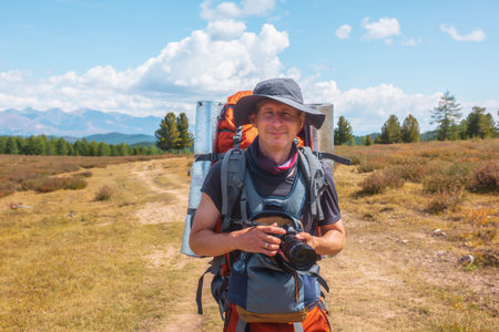 Alone traveler with large backpack walks along hiking trail on sunlit high mountain plateau under white clouds in blue sky. Backpacker with photo camera in autumn mountain trekking in good weather.の写真素材
