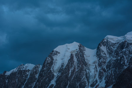 Atmospheric nightly landscape with huge snowy mountain top in dramatic sky. Hanging glacier and cornice on beautiful giant snow mountains in night. High snow-covered mountain range in dusk dim light.の写真素材