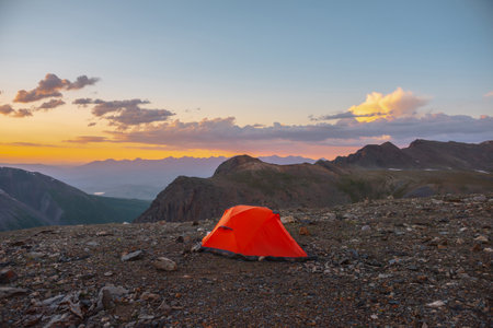 Scenic alpine landscape with tent at very high altitude with view to large mountains in orange dawn sky. Vivid orange tent with awesome view to high mountain range under cloudy sky in sunset colors.の写真素材