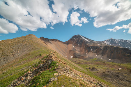 Scenic landscape with sunlit green grassy stone hill and high orange mountain ridge under white clouds in blue sky. Colorful scenery with rocky mountains and sharp rocks in sunlight under cloudy sky.の写真素材