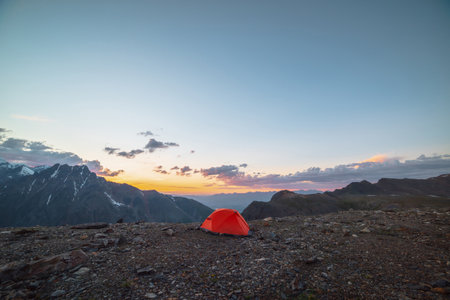 Scenic alpine landscape with tent at very high altitude with view to large mountains in orange dawn sky. Vivid orange tent with awesome view to high mountain range under cloudy sky in sunset colors.の写真素材