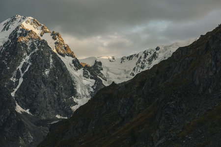 Gloomy landscape with giant snowy mountain range and sunlit gold rocks in dramatic gray sky. Huge snow mountains with hanging glacier and cornice under rainy clouds. Snow-covered mountains in overcastの写真素材