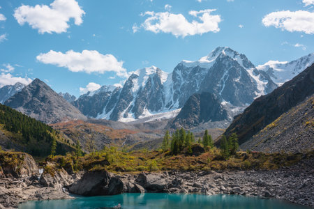 Pure turquoise alpine lake with view to forest hill and giant snow mountains in autumn sunny day. Glacial lake against huge snow covered mountain range in bright sun. Vivid colors in high mountains.の写真素材