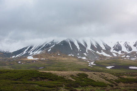 Dramatic alpine landscape with snowy mountains in gray low clouds. Bleak atmospheric scenery of tundra under lead gray sky. Gloomy minimalist view to mountain range among low rainy clouds in overcast.の写真素材
