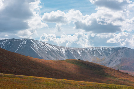 Motley autumn landscape with hills on high plateau and sunlit snowy mountain range under dramatic cloudy sky. Vivid autumn colors in mountains. Sunlight and shadows of clouds in changeable weather.の写真素材