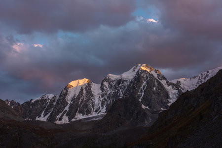 Atmospheric landscape with sunset gold reflection on huge snowy mountain top in violet dramatic sky. Scenic view to giant snow mountains in dusk. Snow-covered mountain range silhouettes in twilight.の写真素材