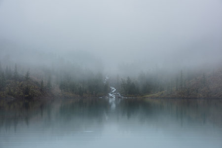 Mountain creek flows from forest hills into glacial lake in mysterious fog. Small river and coniferous trees reflected in calm alpine lake in misty morning. Tranquil landscape in fading autumn colors.の写真素材