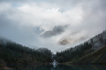 Tranquil scenery with alpine lake. Mountain creek flows from forest hills into lake. Snowy mountains in fog clearance. Small river and coniferous trees in low clouds.の写真素材