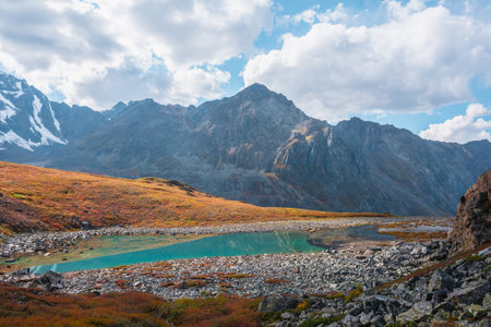 Lovely autumn landscape with turquoise mountain lake against high mountain top in sunlight under clouds in blue sky. Sunlit beautiful small alpine lake and large mountain range. Vivid autumn colors.の写真素材