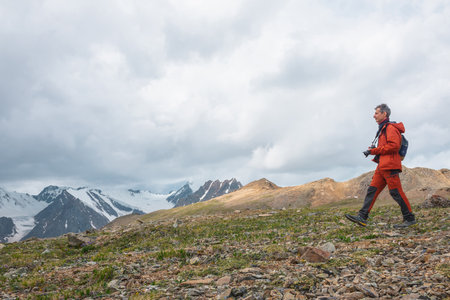 Man in red with camera walks with view to large snowy mountain range with sharp tops under gray cloudy sky. Dramatic landscape with tourist in high mountains. Snow pointy peaks at changeable weather.の写真素材