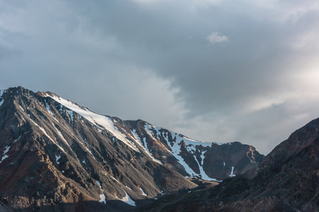 Dramatic overcast landscape with sunlit high mountain range with sharp rocks under gloomy sky. Dark atmospheric mountain scenery with large mountains in sunlight under cloudy sky at changeable weatherの写真素材