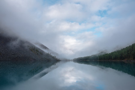 Tranquil meditative misty scenery of glacial lake with reflection of pointy fir tops and clouds at early morning. Mountain lake in fog.の写真素材