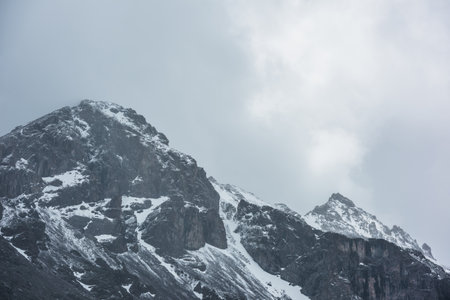 Awesome landscape with high snowy mountain peaked top with sharp rocks in cloudy sky. Dramatic view to snow mountain pointed peak in rainy weather. Atmospheric scenery with white snow on black rocks.の写真素材