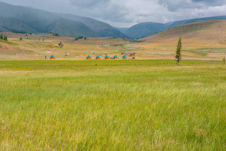 Scenic view from sunlit grassy steppe to small houses on background of green high mountain range in rainy clouds. Dramatic landscape with meadow and small lodges in mountains at changeable weather.の写真素材