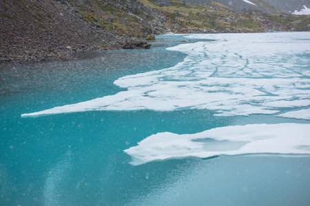 Atmospheric mountain landscape with frozen alpine lake during snowfall. Clear ice floats on water surface of lake. Awesome mountain scenery with icy mountain lake. Flakes of snow on water background.の写真素材