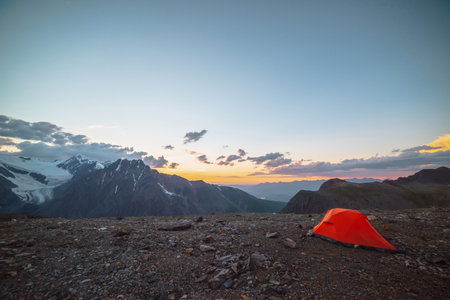 Scenic alpine landscape with tent at very high altitude with view to large mountains in orange dawn sky. Vivid orange tent with awesome view to high mountain range under cloudy sky in sunset colors.の写真素材