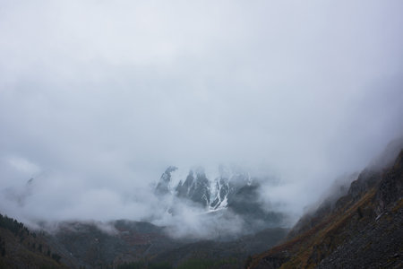 Dark atmospheric landscape with high snow mountains in dense fog in rainy weather. Large snowy mountain range in thick fog in dramatic overcast. Snow-covered rocky mountains in low clouds during rain.の写真素材