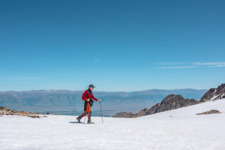 Tourist in red walks on snow mountain near abyss edge on high altitude under blue sky in sunny day. Man with camera on snowy mountain near precipice edge with view to large mountain range in away.の写真素材