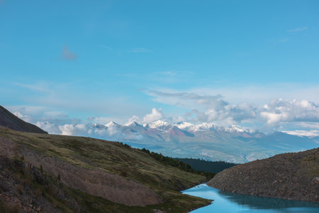 Atmospheric landscape with azure mountain lake against high snow-white mountain range in sunlight in low clouds. Dramatic evening view to alpine lake and large sunlit snow mountains in low clouds.の写真素材