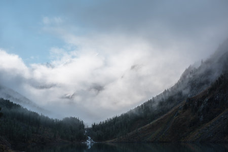 Tranquil scenery with snow mountain range in clouds and alpine lake. Mountain creek flows from forest hills into glacial lake. Snowy mountains in fog clearance. Small river and trees in low clouds.の写真素材