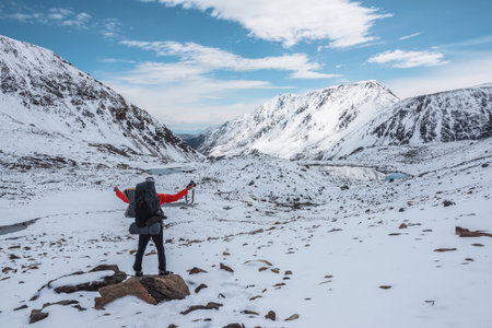 Man in red jacket with big backpack in snowy valley with alpine lakes under clouds in blue sky. Backpacker in snow-covered large mountains among glacial lakes, pure white snow and stones in sunny day.の写真素材