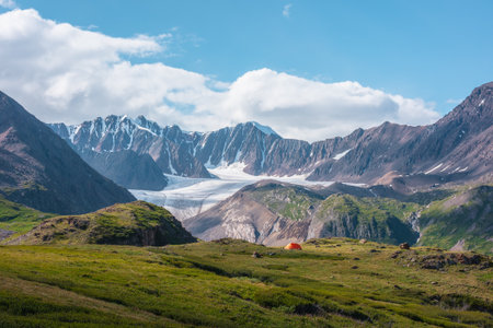 Colorful landscape with orange tent in alpine valley with view to big glacier tongue and large snow-capped mountain range under clouds in blue sky. Green hills and rocks against ice and sheer crags.の写真素材