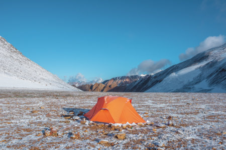 Orange tent on snow-covered stony pass in sunlight. Snowy stone hills and rocky mountain ridge at early morning. Red tent in high mountains in freshly fallen snow. Low clouds in blue sky at sunrise.の写真素材