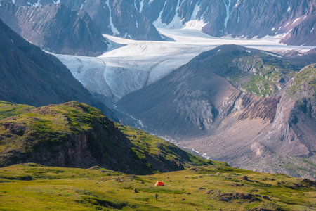 Vivid orange tent in sunlit vast alpine valley among green hills and rocks with view to big glacier tongue and large rocky wall in sunny day. Awesome colorful scenery with ice and sheer crags close upの写真素材