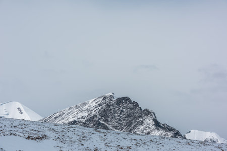 Sunlit snowy rocky pointy peak in gray cloudy sky. Snow-covered sharp rocky mountain range and...