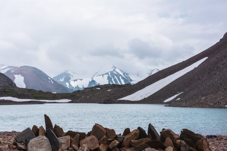 Sharp pointed stones on background azure alpine lake against snow-covered few pointy peaks in low clouds. Ripples on blue water surface of mountain lake and three snowy peaked tops in low cloudiness.の写真素材