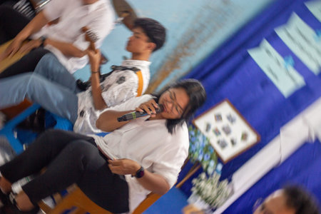 Muntilan, Indonesia - 16 Mar 2026: Woman sitting and singing passionately into a microphone during a school event or talent show performance, with students in the backgroundのeditorial素材