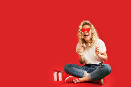 beautiful young girl with red hearts and a white gift with a bow sits in full growth on an isolated...