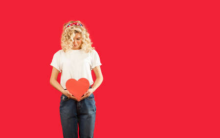 beautiful young emotional girl with a red heart in her hands is standing on an isolated red...