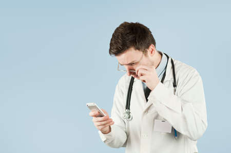 Young cute doctor student with a smartphone in his hands talking to friends on a blue isolated background. Electric conflict concept and connectionの写真素材