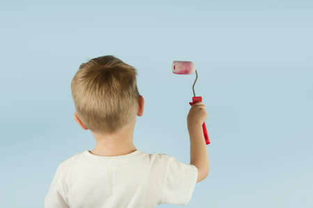 Beautiful blond child boy paints the wall with a roller and writes paint on the wall on an isolated blue background. Concept of discounts and salesの写真素材