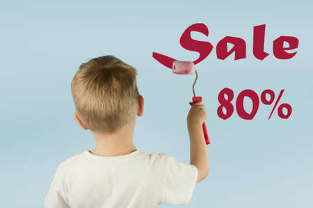 Beautiful blond child boy paints the wall with a roller and writes paint on the wall on an isolated blue background. Concept of discounts and salesの写真素材