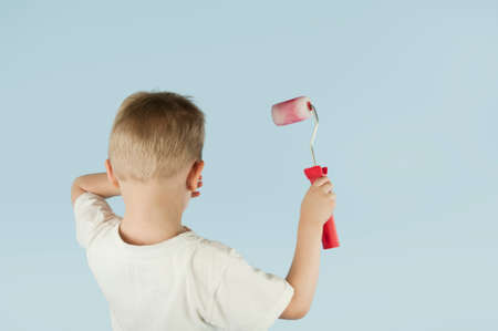 Beautiful blond child boy paints the wall with a roller and writes paint on the wall on an isolated blue background. Concept of discounts and salesの写真素材