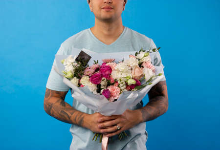 Unrecognizable man in blue hirt holding flowers as gift for his mother or girlfriend on birthday. Anniversary present concept. Studio shot on colored wall.の写真素材