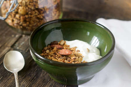 A useful breakfast is yoghurt with granola. Granola with yoghurt in a ceramic bowl and granola in a glass jar on a wooden background. Rustic style, selective focus.の写真素材