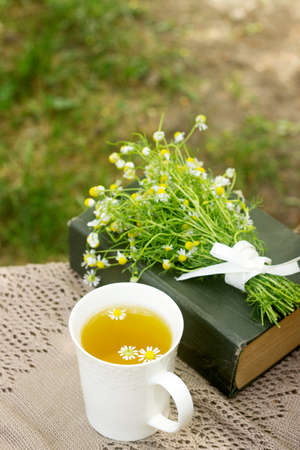 A cup of useful soothing chamomile tea, a bouquet of chamomiles and a book on a swing. Selective focus.の写真素材