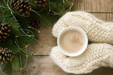 Women's hands in mittens hug a cup of cocoa on the background of fir branches with pine cones and a...
