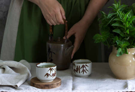 Mint tea in a vintage Chinese tea set, a woman in a linen dress and an apron, and a bouquet of mint in a jug.の写真素材