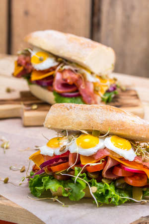 A sandwich with bacon, cheese and fried quail eggs. A sandwich with fresh vegetables and herbs on a wooden background. Rustic.の写真素材