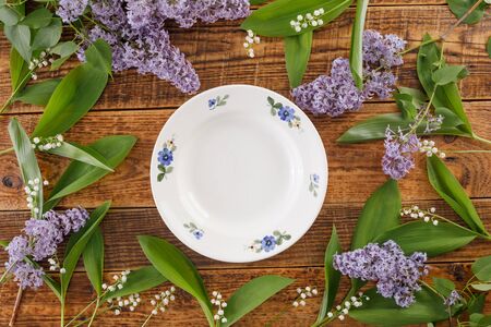 Plate and decor of flowers of lilies of the valley and lilac on the background of vintage wooden boards. Vintage background with flowers and place under the text. View from above. Flat lay. Cutlery. Vintage.の写真素材