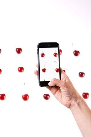 Smartphone in hand. Produces photos of sweet cherry berries on a white background. Juicy summer berries closeup.Isolated.の写真素材