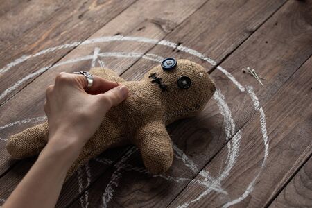 Voodoo doll on a wooden background with dramatic lighting. The concept of witchcraft and black art and the occult. Burlap doll on the background of a drawn star. Hand sticking pins into a doll. Copy space.の写真素材