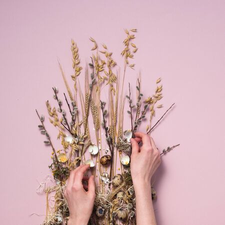 Female hands collect easter composition on a pale pink background. Dried flowers and eggs for a beautiful greeting card. The concept of Easter decorative cards. Square.の写真素材