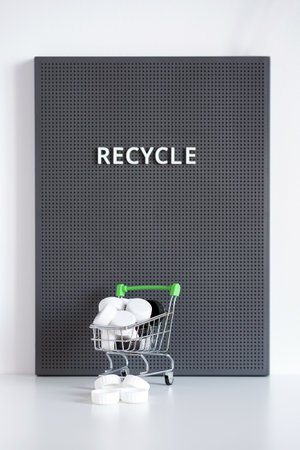 Black and white lids in a small shopping cart on a grey board with recycle word. Recycling concept....